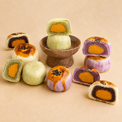 A group of colorful Chinese sweet pastries, including whole and sliced mooncakes, arranged on a beige background. The pastries feature various fillings such as salted egg yolk, taro, and matcha, with some topped with sesame seeds.